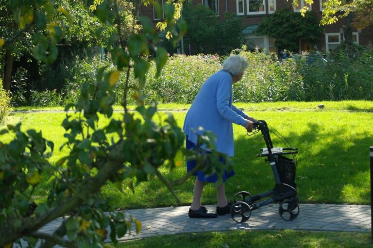 Oudere vrouw met rollator in een zonnig park, omringd door gras en bomen.