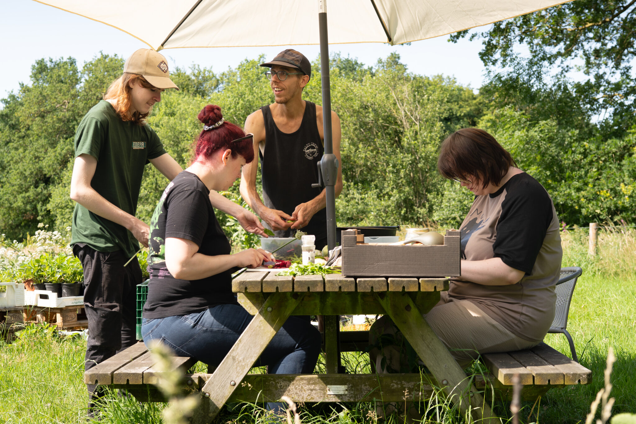 Groep mensen bereidt eten voor op een picknicktafel in een zonnige tuin met bomen op de achtergrond.