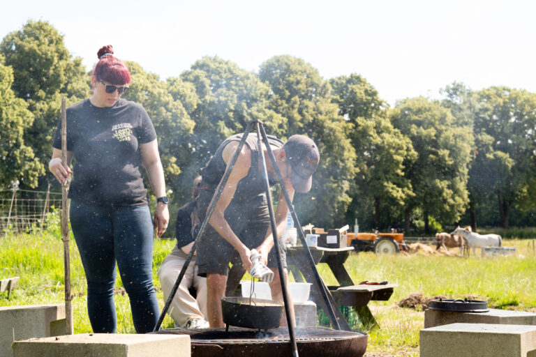 Twee mensen koken buiten bij een vuurpot in een groene, landelijke omgeving met bomen.