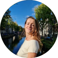 Jonge vrouw glimlacht bij kanaal met bomen en huizen op achtergrond.