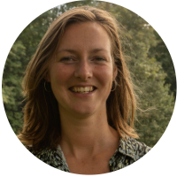 Een vrouw glimlacht in een groene omgeving met bomen op de achtergrond.