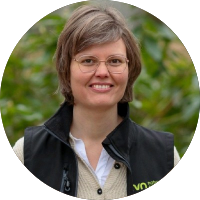 Een persoon met een bril glimlacht buiten, omgeven door groen.