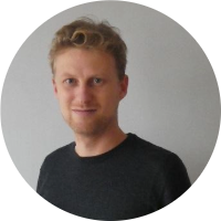 Een man met blond haar, glimlachend en gekleed in een donker shirt tegen een grijze achtergrond.