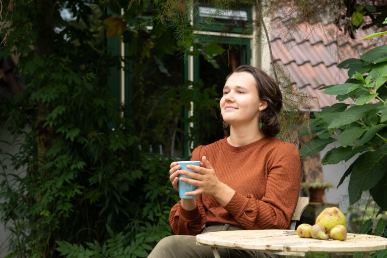 Persoon in bruine trui drinkt koffie in een tuin, zittend aan tafel met peren erop.