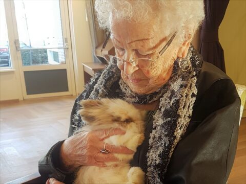 Oudere vrouw met bril knuffelt een zacht konijn in een lichte kamer.
