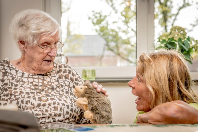 Twee vrouwen delen een moment met een pluchen egeltje bij een raam.