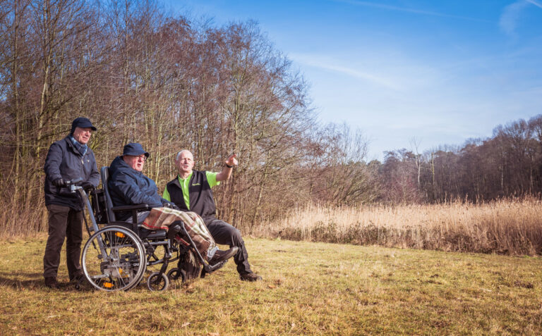 Drie mannen genieten van een zonnige dag in de natuur; een zit in een rolstoel.