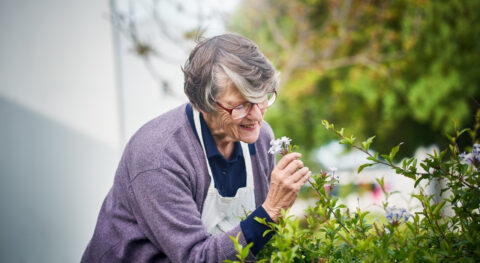 Oudere vrouw geniet van bloemen in een tuin. Ze draagt een bril en een paarse trui.
