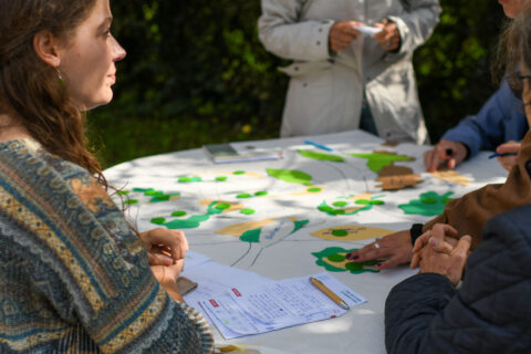 Mensen aan tafel overleggen met kaarten en notities, buiten in daglicht.