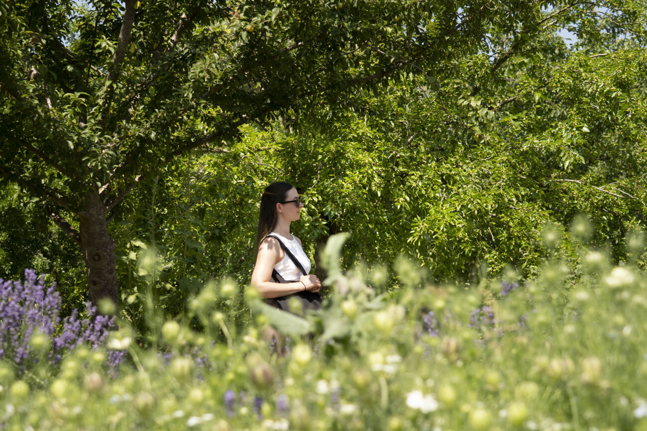Vrouw met zonnebril staat in een groene tuin omringd door bloemen.