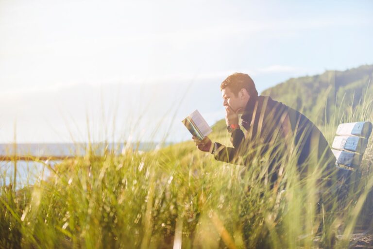 Man leest een boek op een bankje tussen gras, met zee en heuvels op de achtergrond.