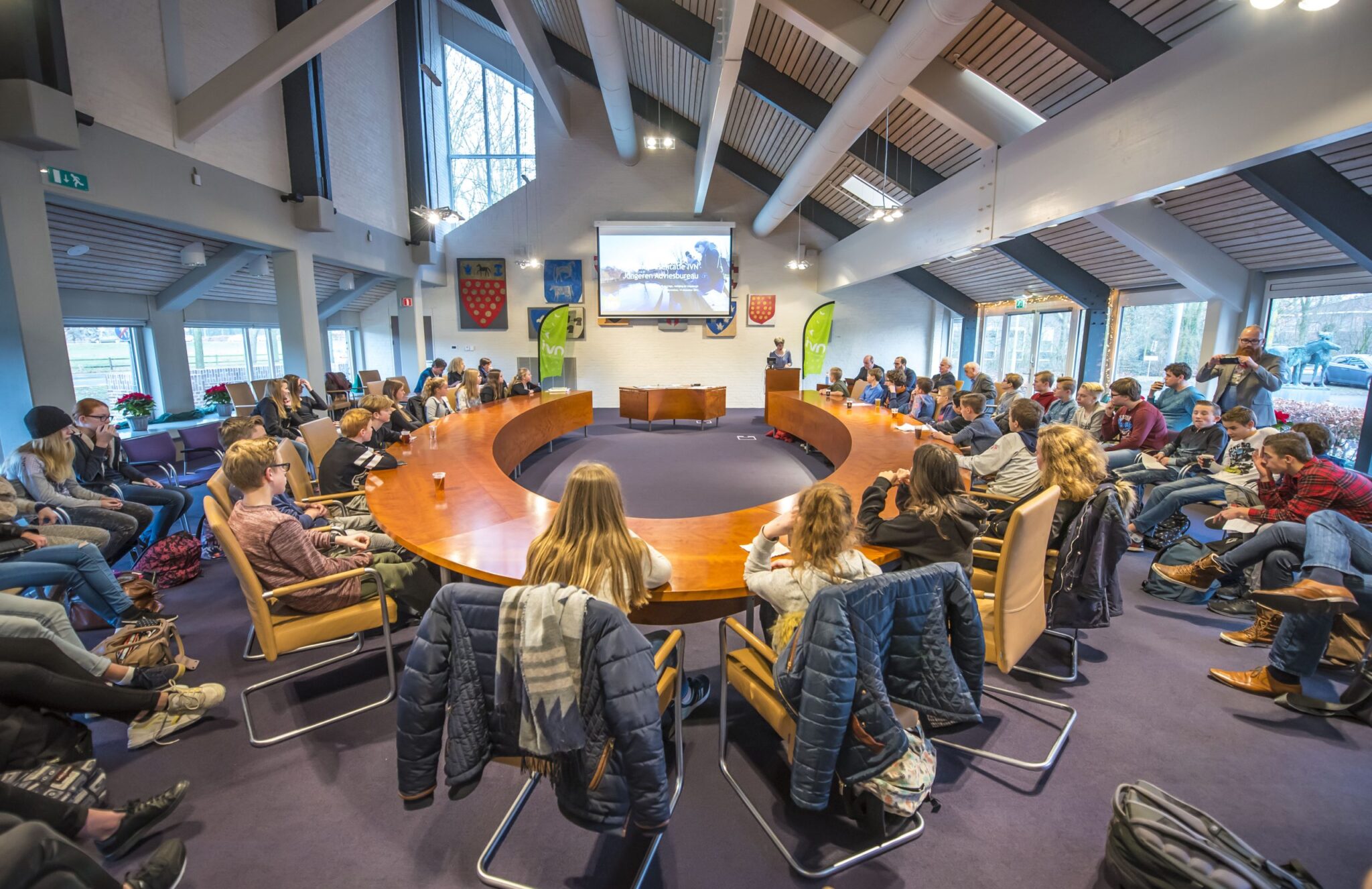 Ronde vergadertafel met mensen die naar een presentatie kijken in een lichte, moderne ruimte.