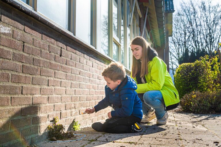 Kind tekent met krijt op een bakstenen muur, vrouw kijkt toe. Buiten, zonnige dag.