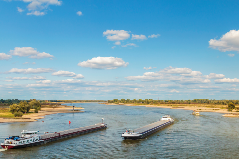 Rivier met twee boten, omgeven door bomen en blauwe lucht met wolken.
