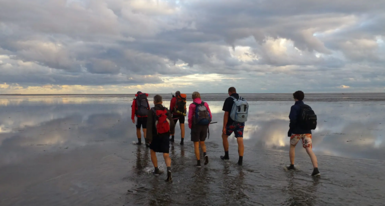 Groep wandelaars met rugzakken loopt over een natte wadvlakte bij bewolkte lucht.