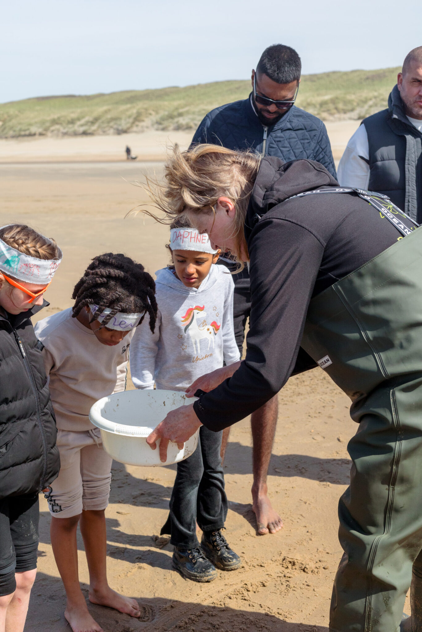 Kinderen kijken samen met een volwassene in een witte kom op het strand.