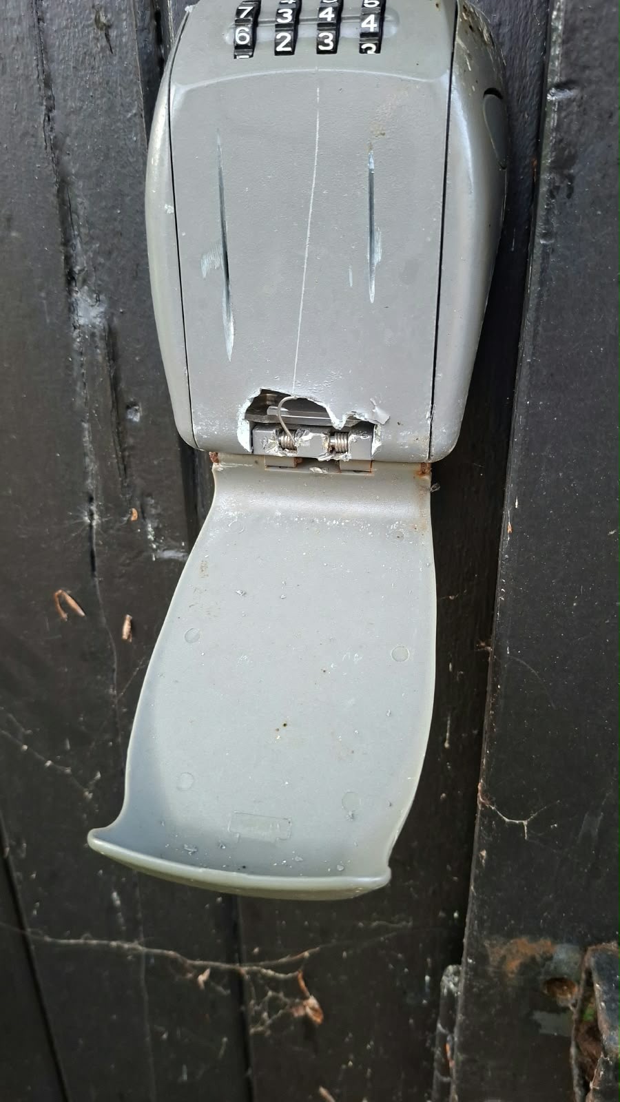 Beschadigde grijze sleutelkluis met cijfercombinatie, openstaand op een donkere houten achtergrond.