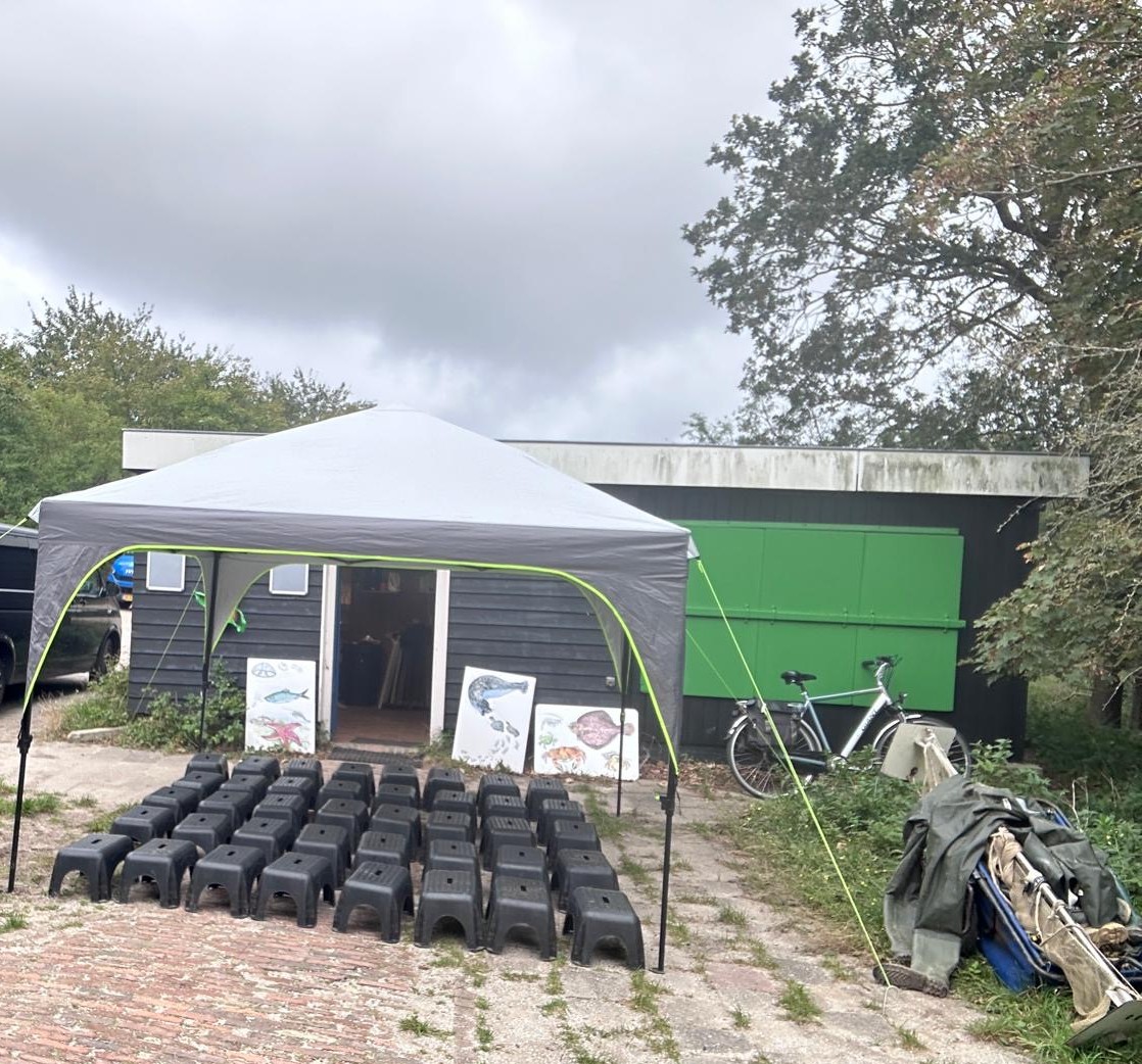Buitentent met zwarte krukjes ervoor, posters van vissen en een fiets naast een schuur.