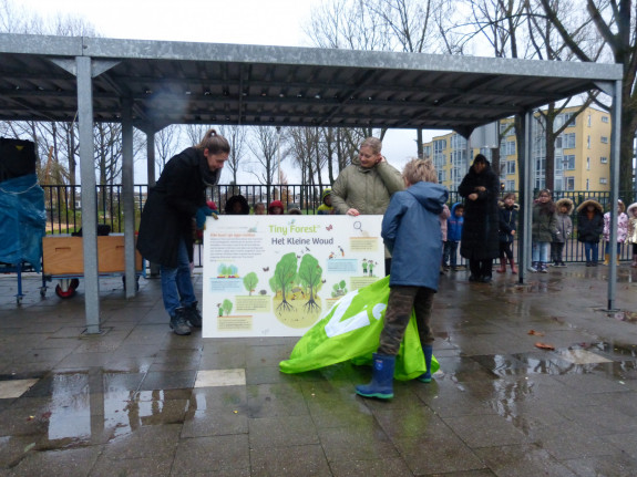 De leerling die de naam voor het Tiny Forest heeft bedacht onthuld het Tiny Forest informatiebord. Zijn moeder en klimaatburgemeester kijken naar het bord waarop staat Tiny Forest Het Kleine Woud.