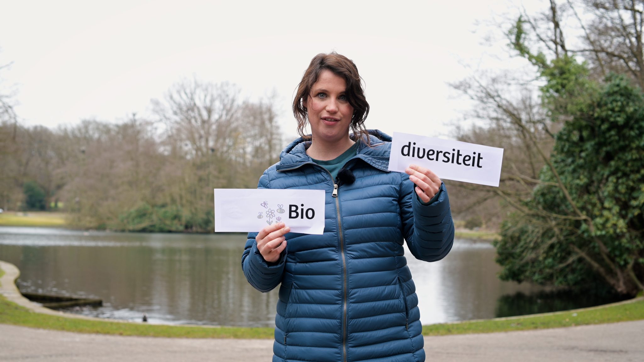 Persoon in blauwe jas houdt borden omhoog met "Bio" en "diversiteit" bij vijver in park.