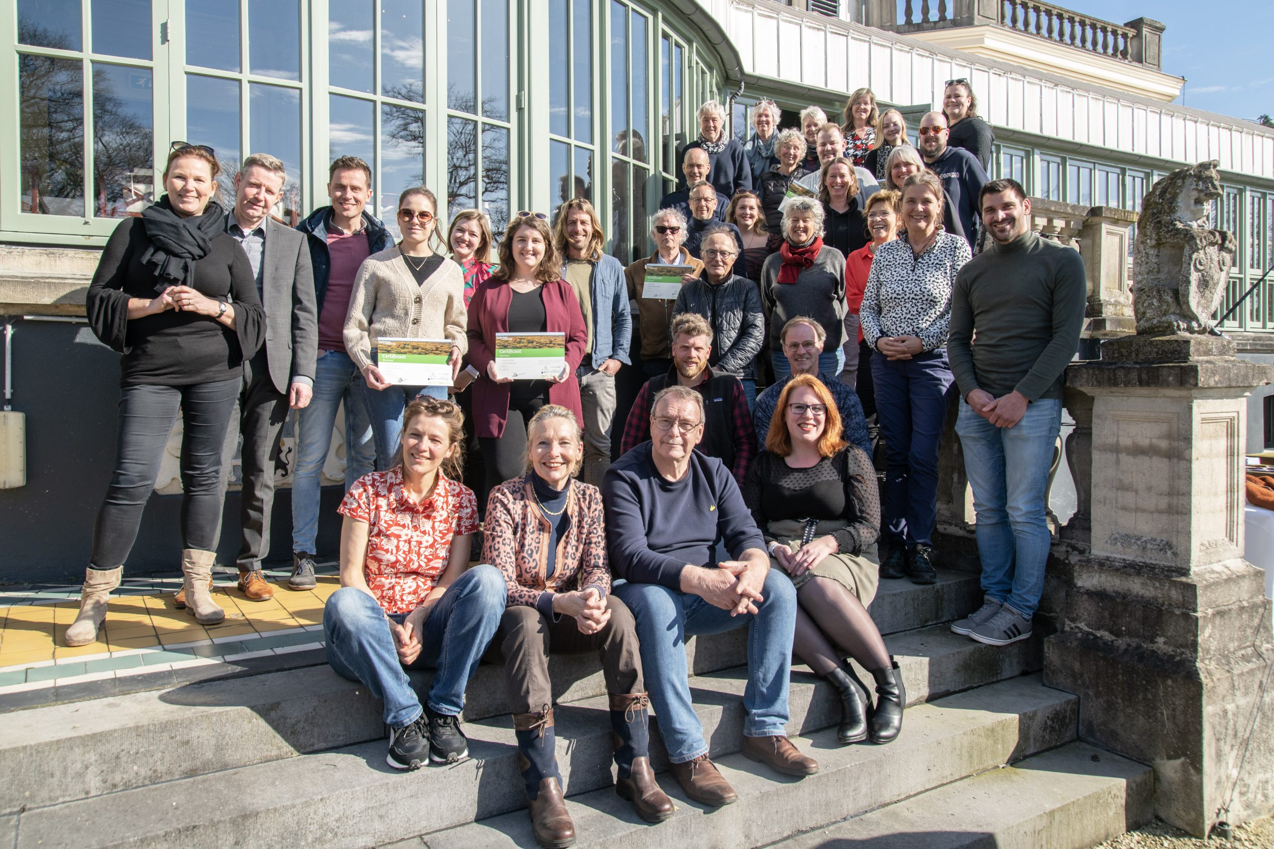 Groepsfoto van mensen op trappen voor een gebouw met glazen gevel en zonnig weer.