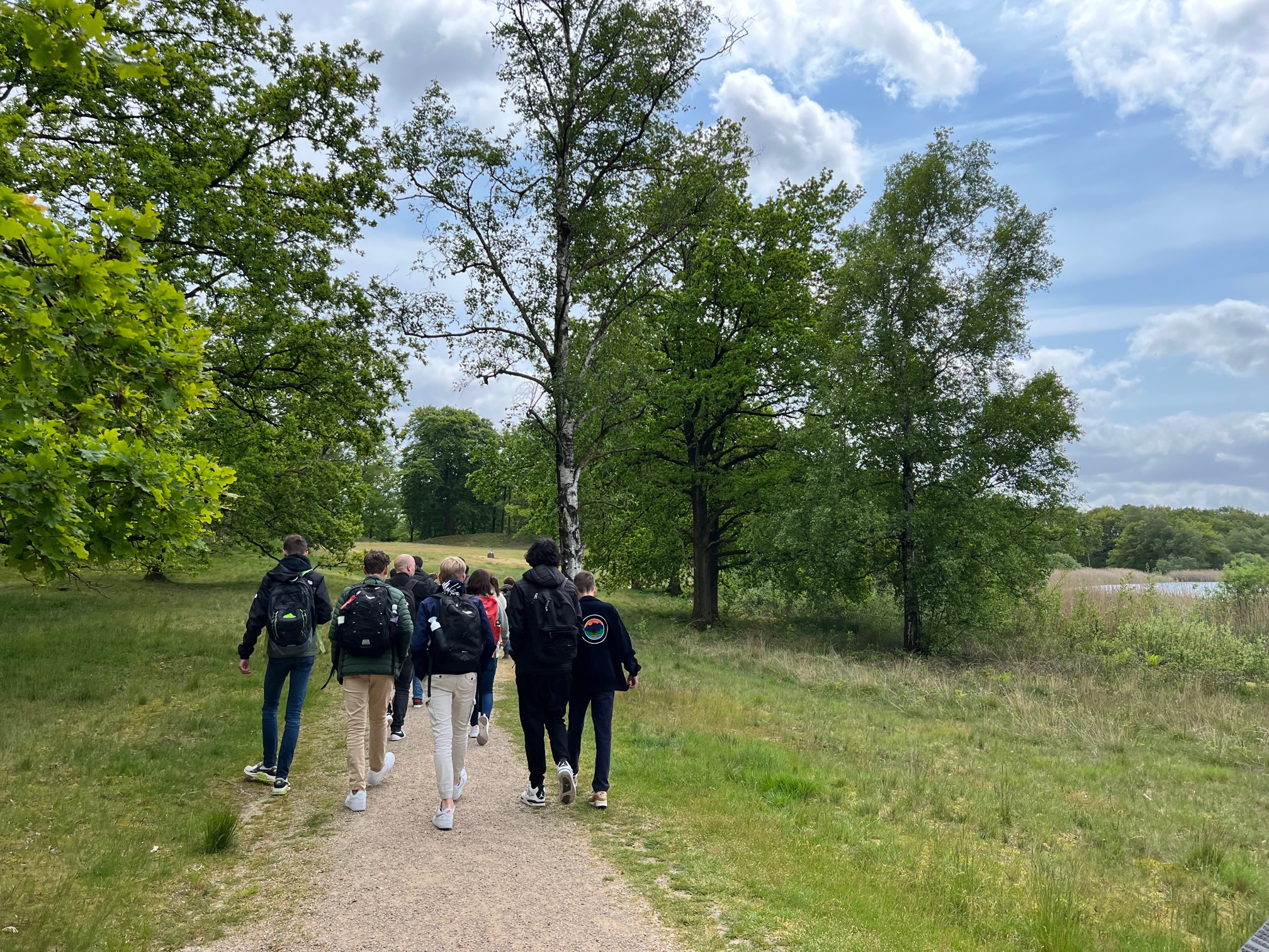 Een groep mensen wandelt op een bospad, omgeven door groene bomen onder een bewolkte hemel.