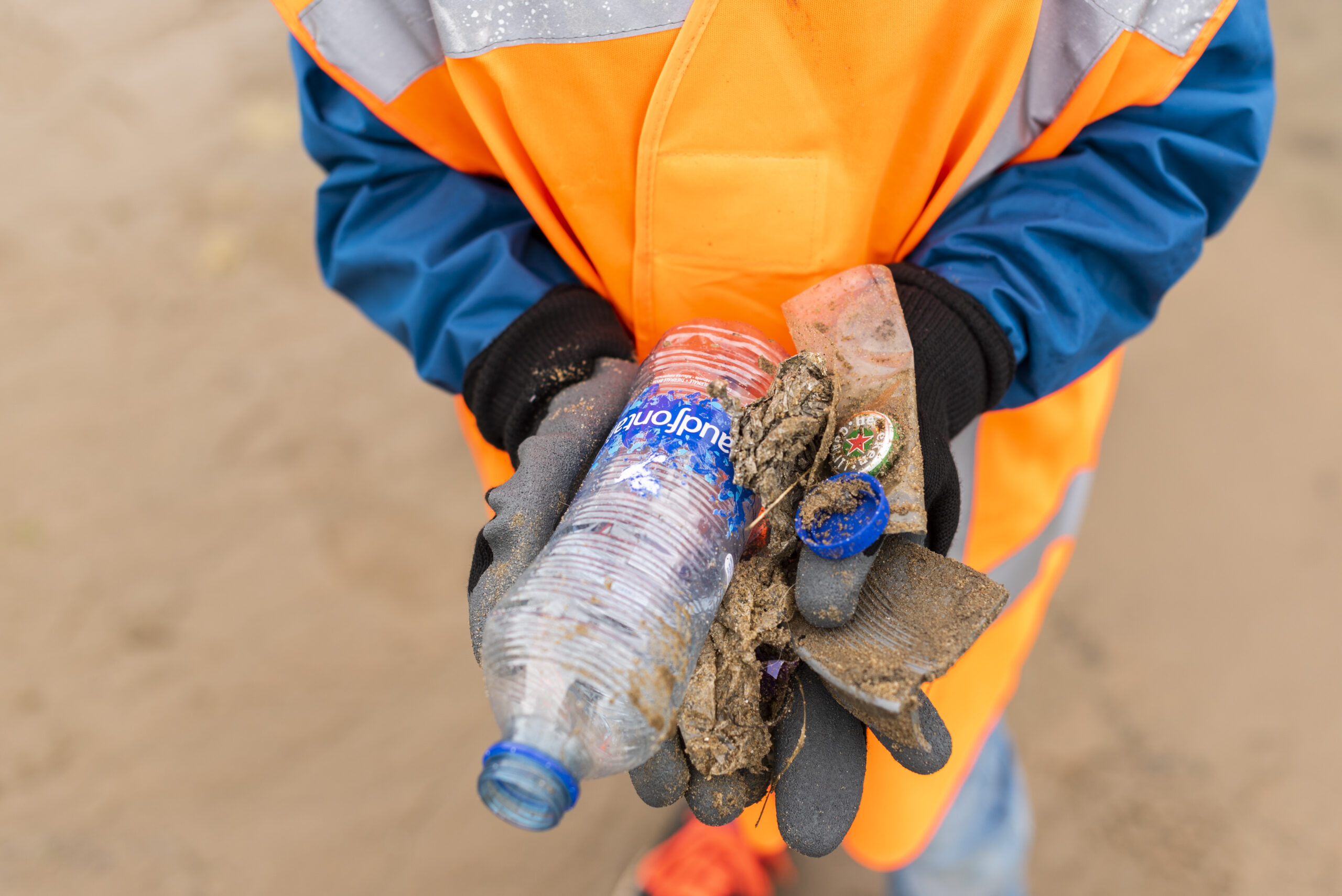 Persoon in oranje vest houdt afval, zoals plastic fles en doppen, in handen op een strand.