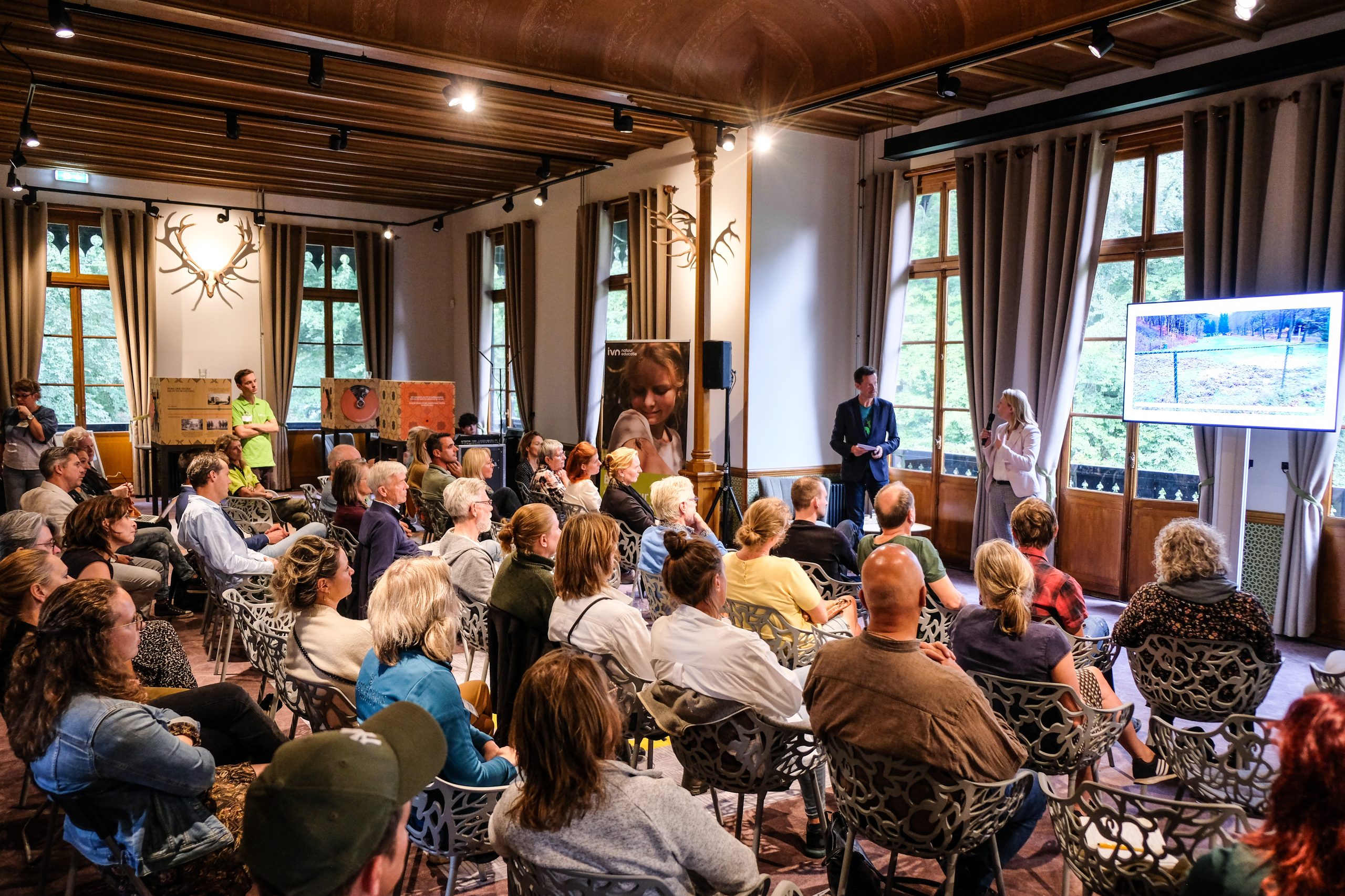 Gepassioneerd publiek luistert naar presentatie met visuals in een zaal met houten details.