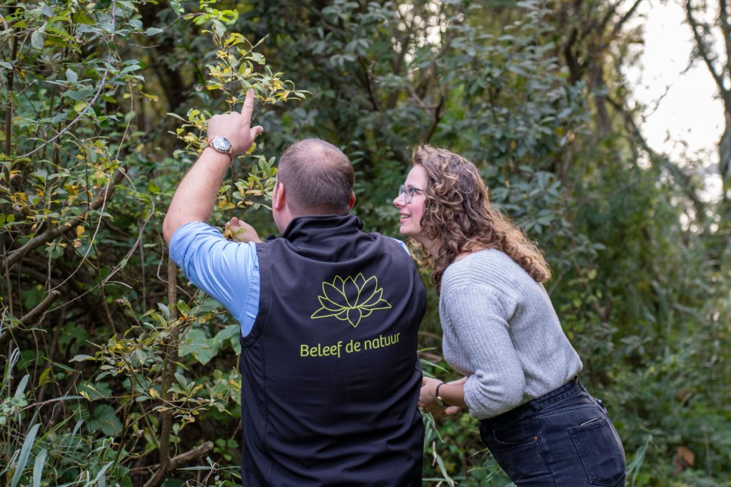 Man en vrouw bestuderen bladeren in bos; man draagt "Beleef de natuur" jas.