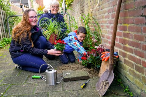 Drie mensen planten bloemen in een tuin, met een gieter en gereedschap op de grond.