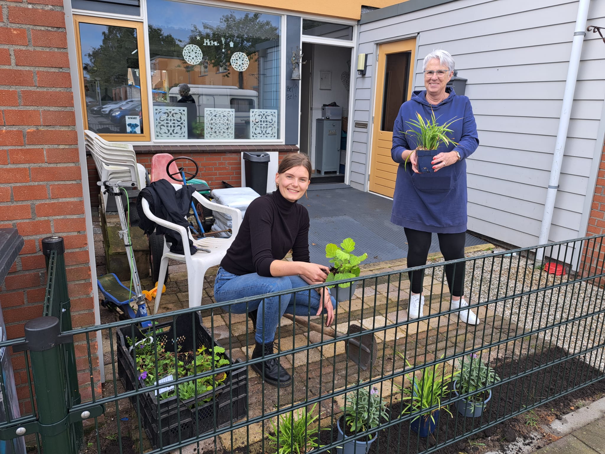 Twee personen planten bloemen in een tuin voor een huis.