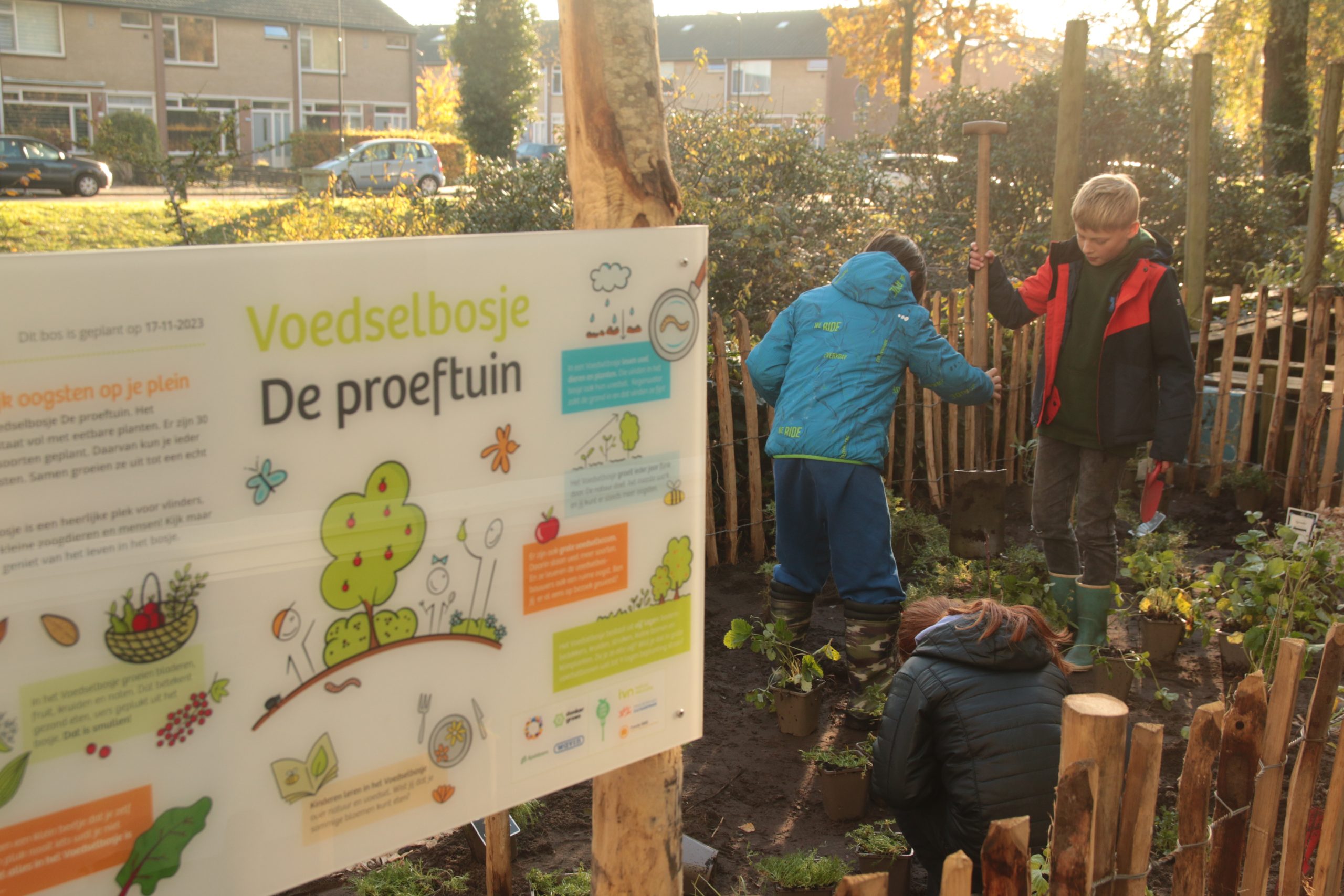 Kinderen werken in een gemeenschapstuin bij een bord met de tekst "Voedselbosje De proeftuin".