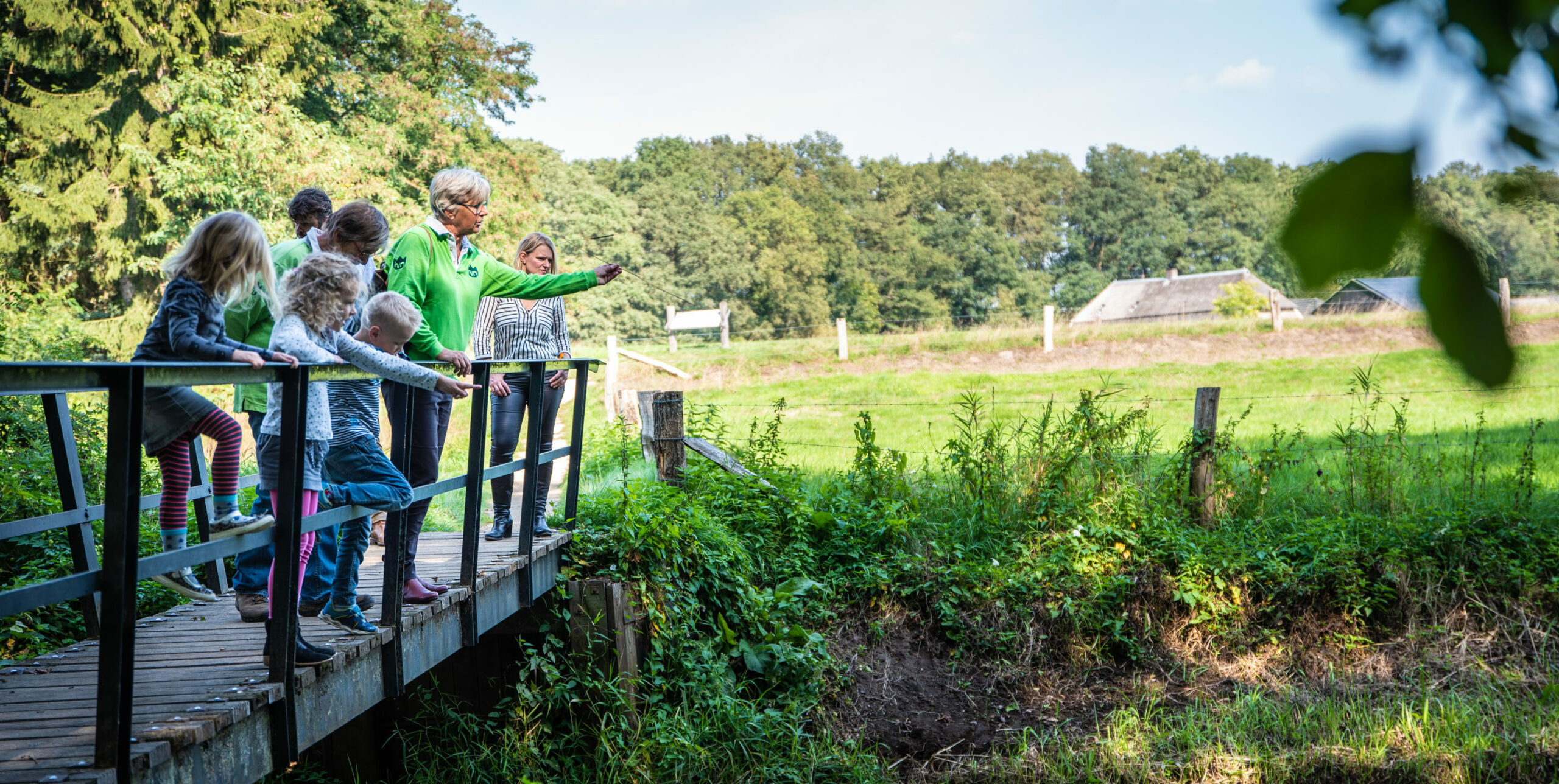 Kinderen en volwassenen op een brug in een groene omgeving, wijzend naar iets in de verte.