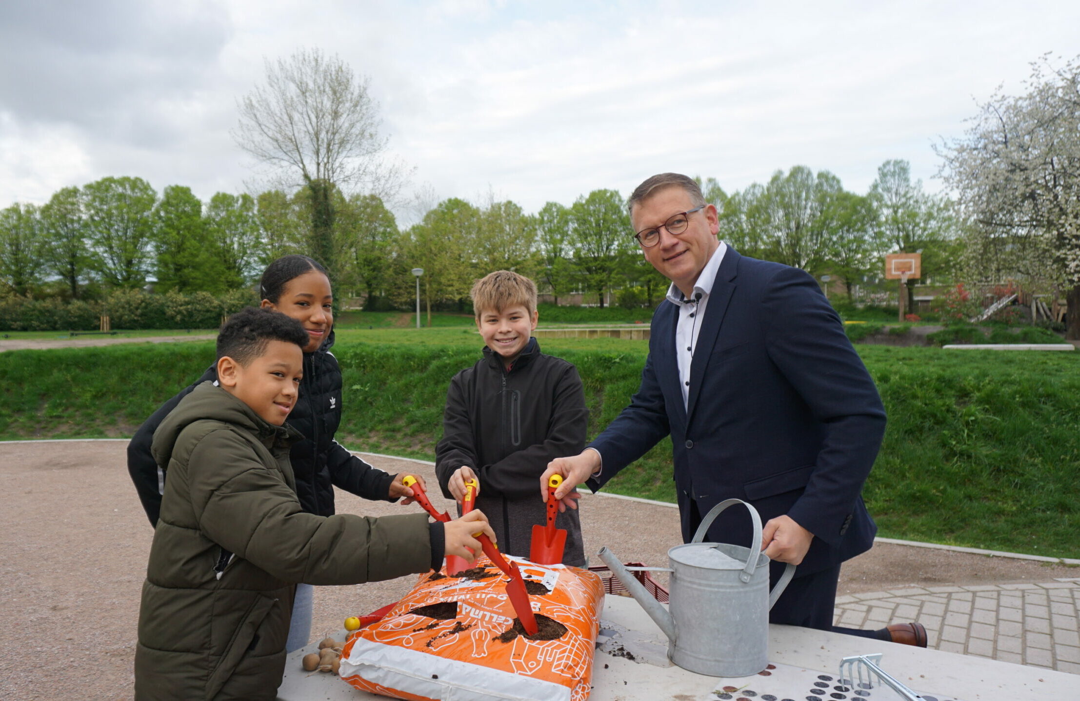 Vier mensen planten zaden met schepjes in een park, met tuinaarde en gieter op tafel.