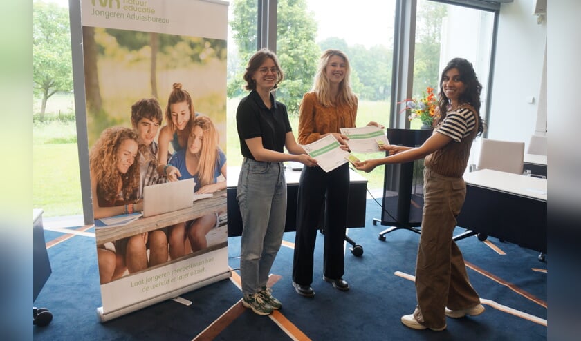 Drie mensen ontvangen certificaten in een kantoor, met een banner en grote ramen op de achtergrond.