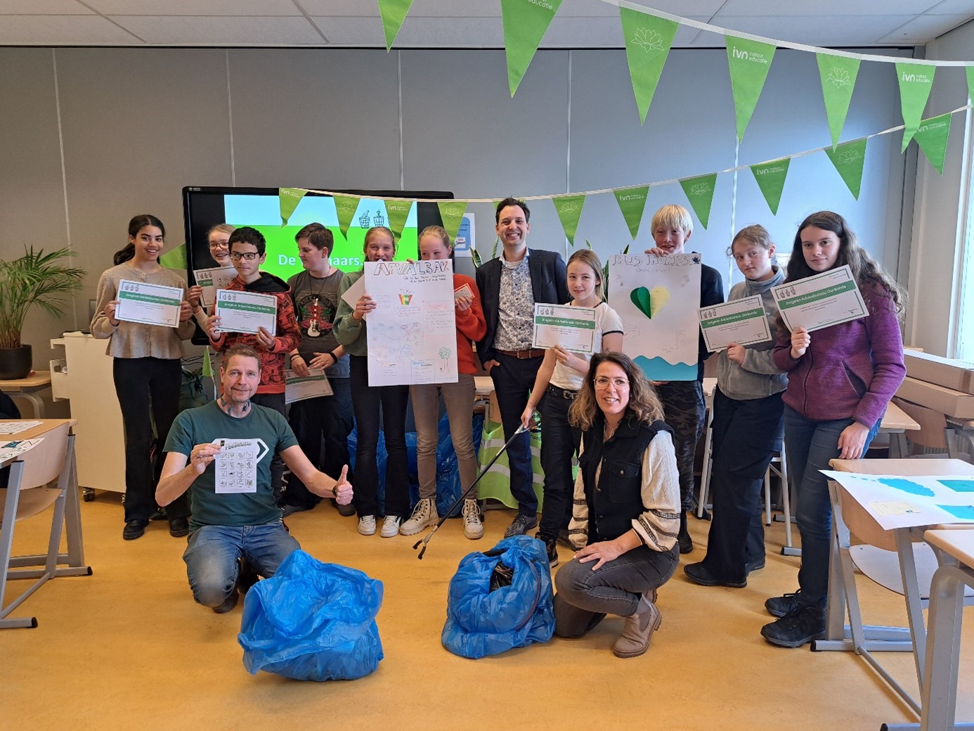 Groep mensen in een klaslokaal met certificaten en milieuposters, omgeven door groene vlaggetjes.