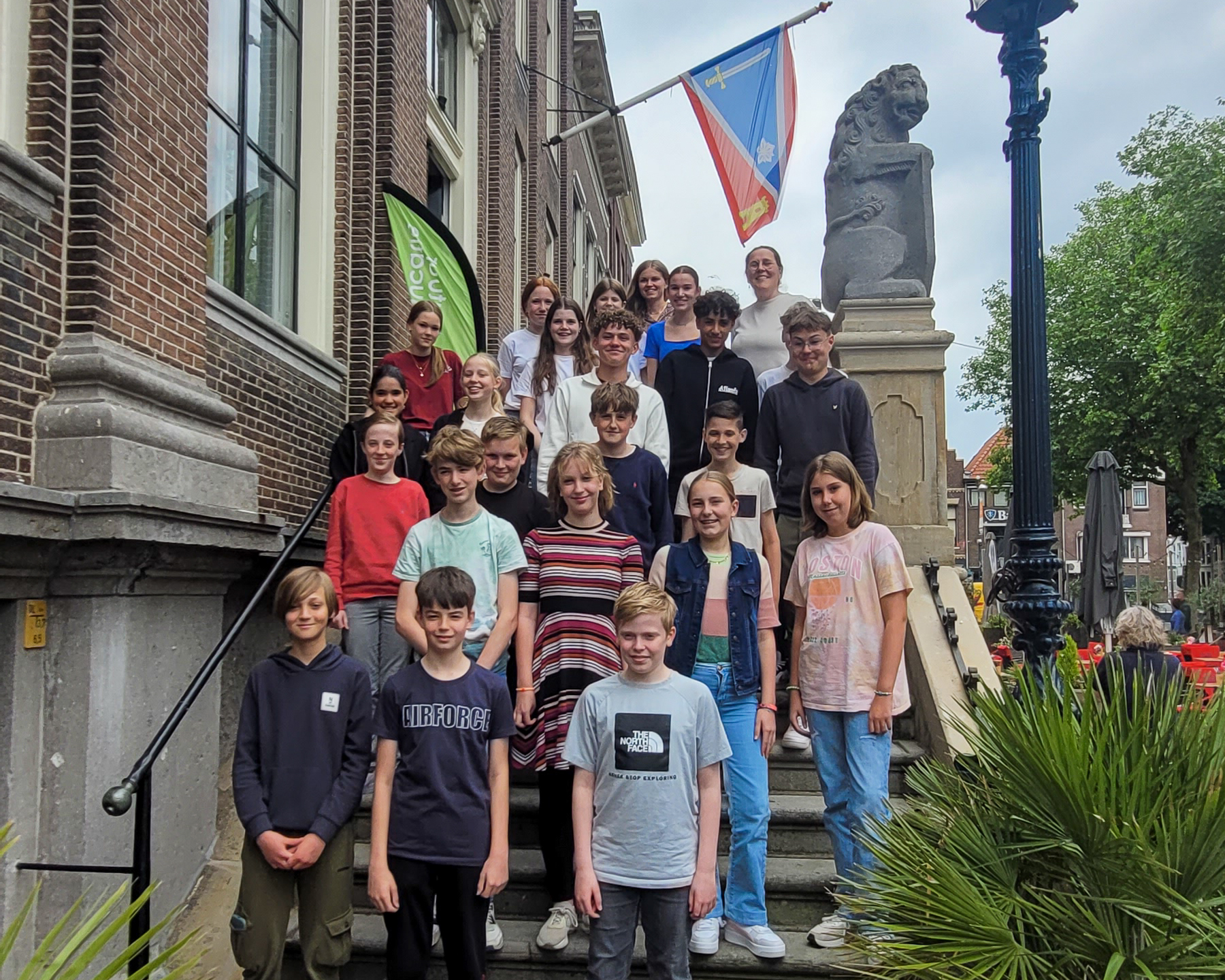 Groep tieners poseert op trap bij historisch gebouw met vlag en leeuwenstandbeeld.