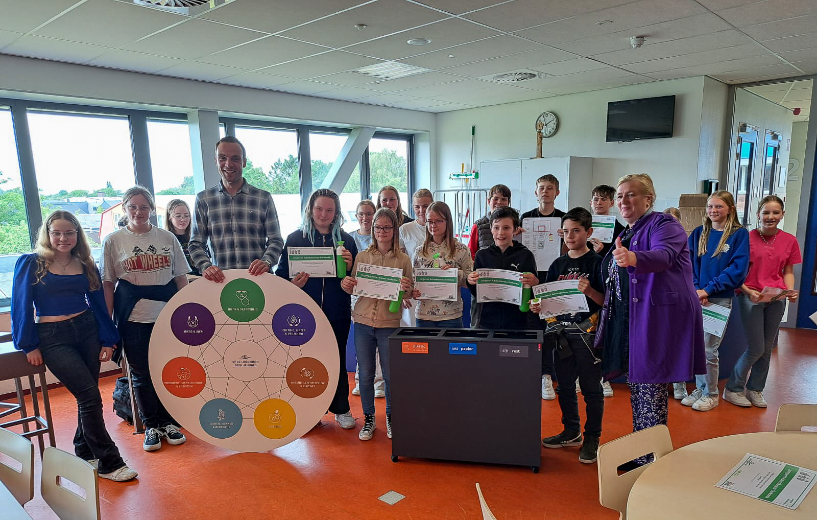 Groep jongeren met certificaten en een infographic over duurzaamheid, naast een recyclebak.