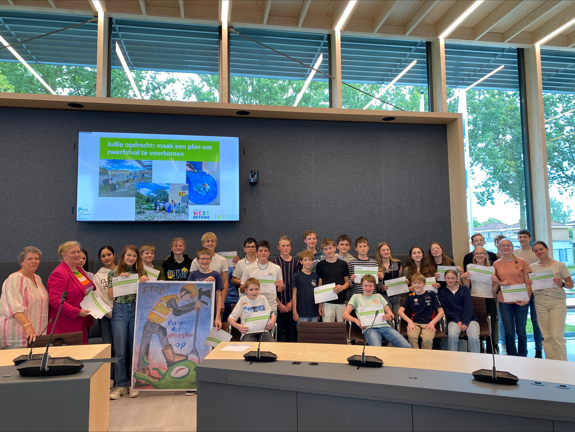 Groep studenten en volwassenen poseren met certificaten in vergaderruimte, dia over zwerfafval.