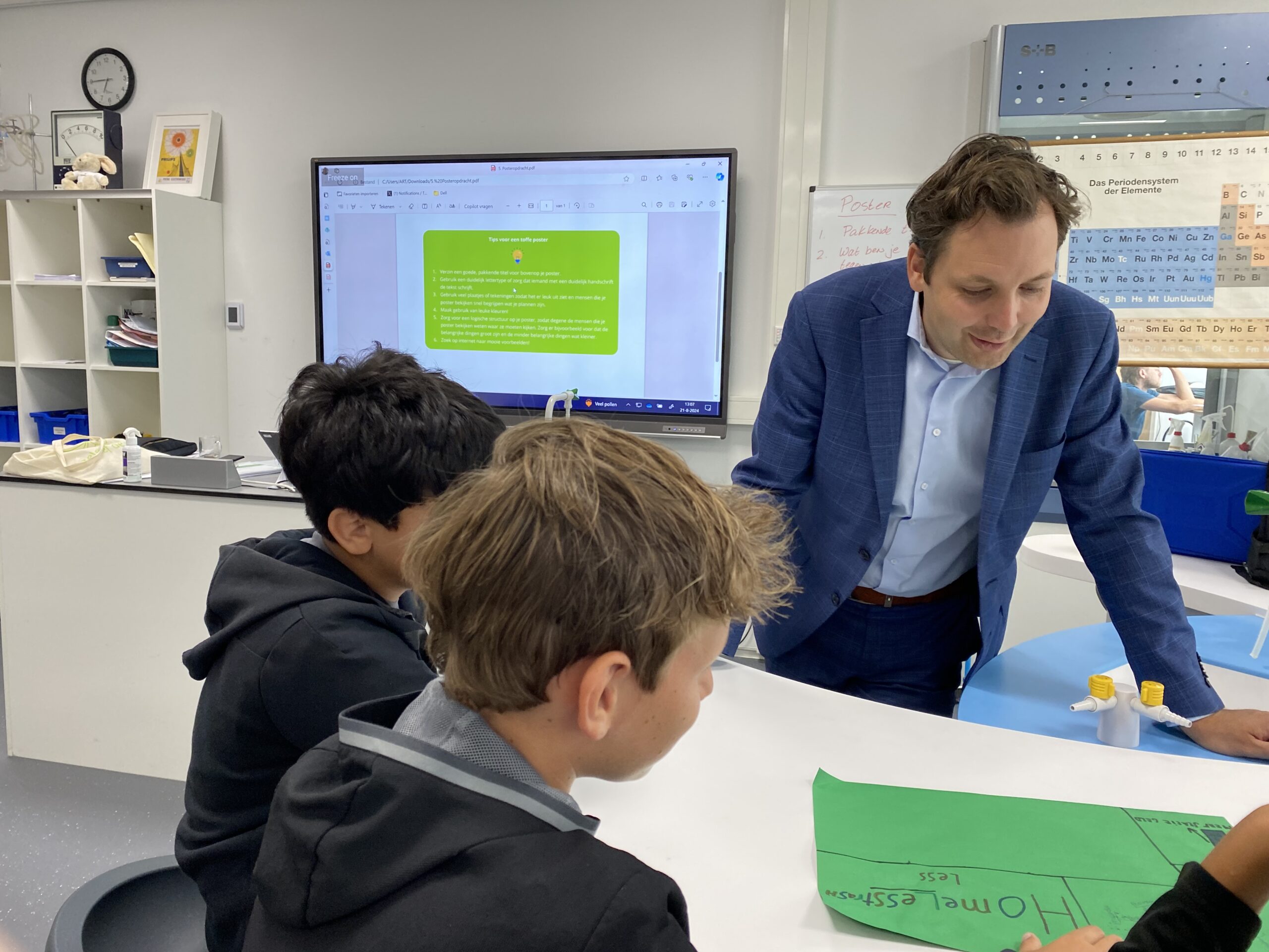 Docent in gesprek met twee leerlingen in een klaslokaal met periodiek systeem aan de muur.