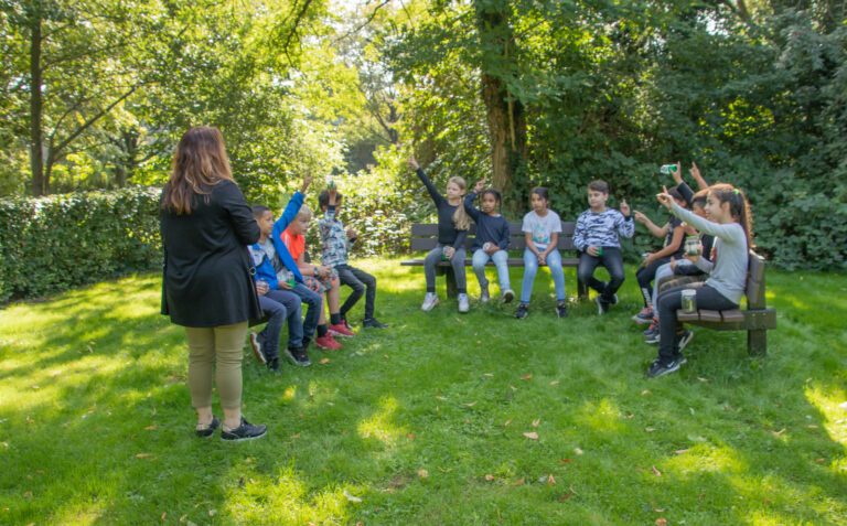 Groep kinderen zit in een cirkel op houten bankjes in een park; een volwassene staat erbij.