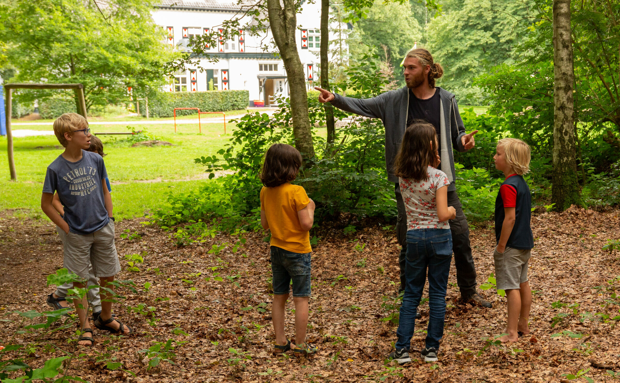 Man wijst iets aan een groep kinderen in een bosrijke omgeving. Op de achtergrond staat een gebouw.
