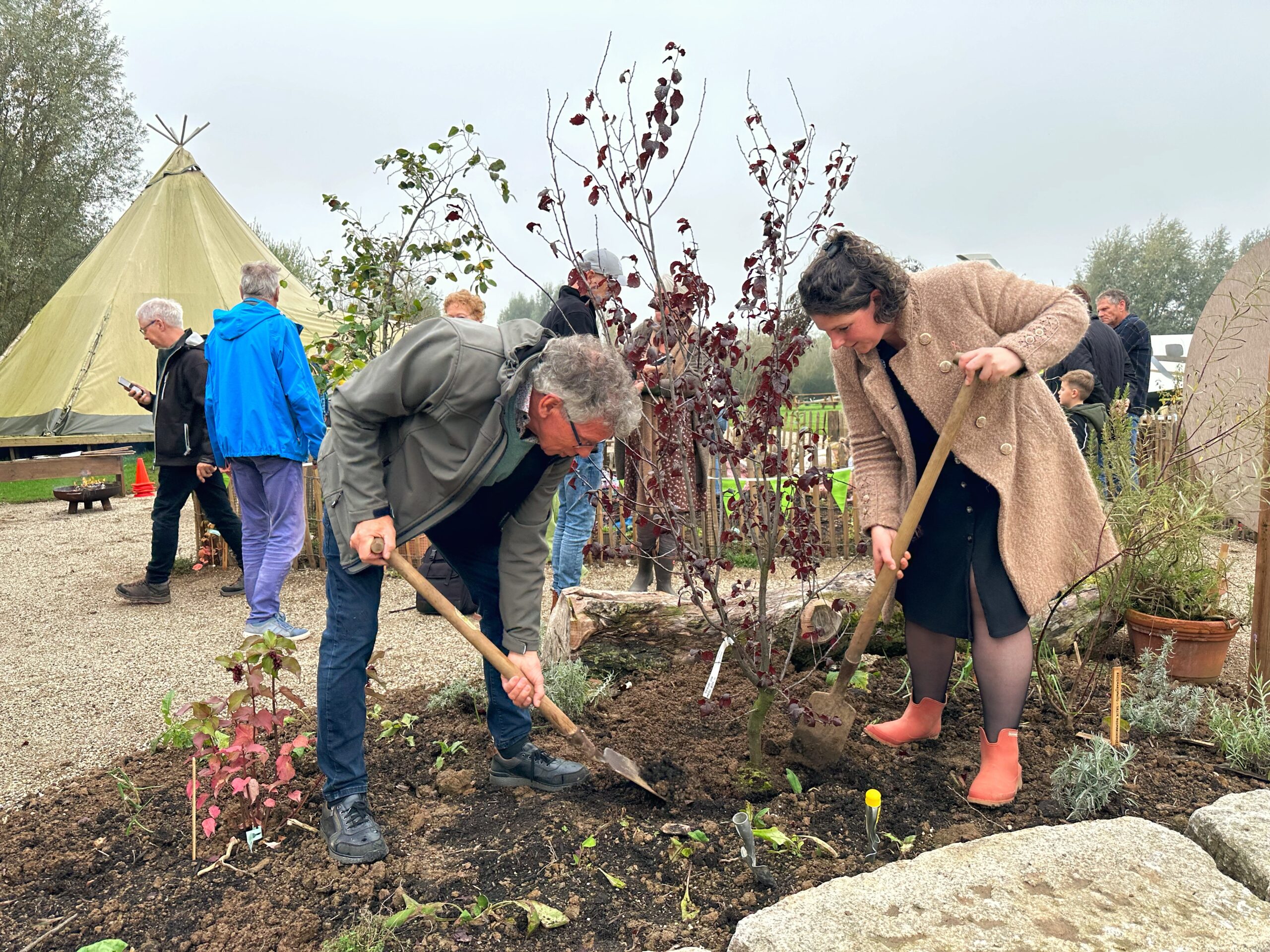 Twee mensen planten een boom naast een tipi, met omstanders op de achtergrond.