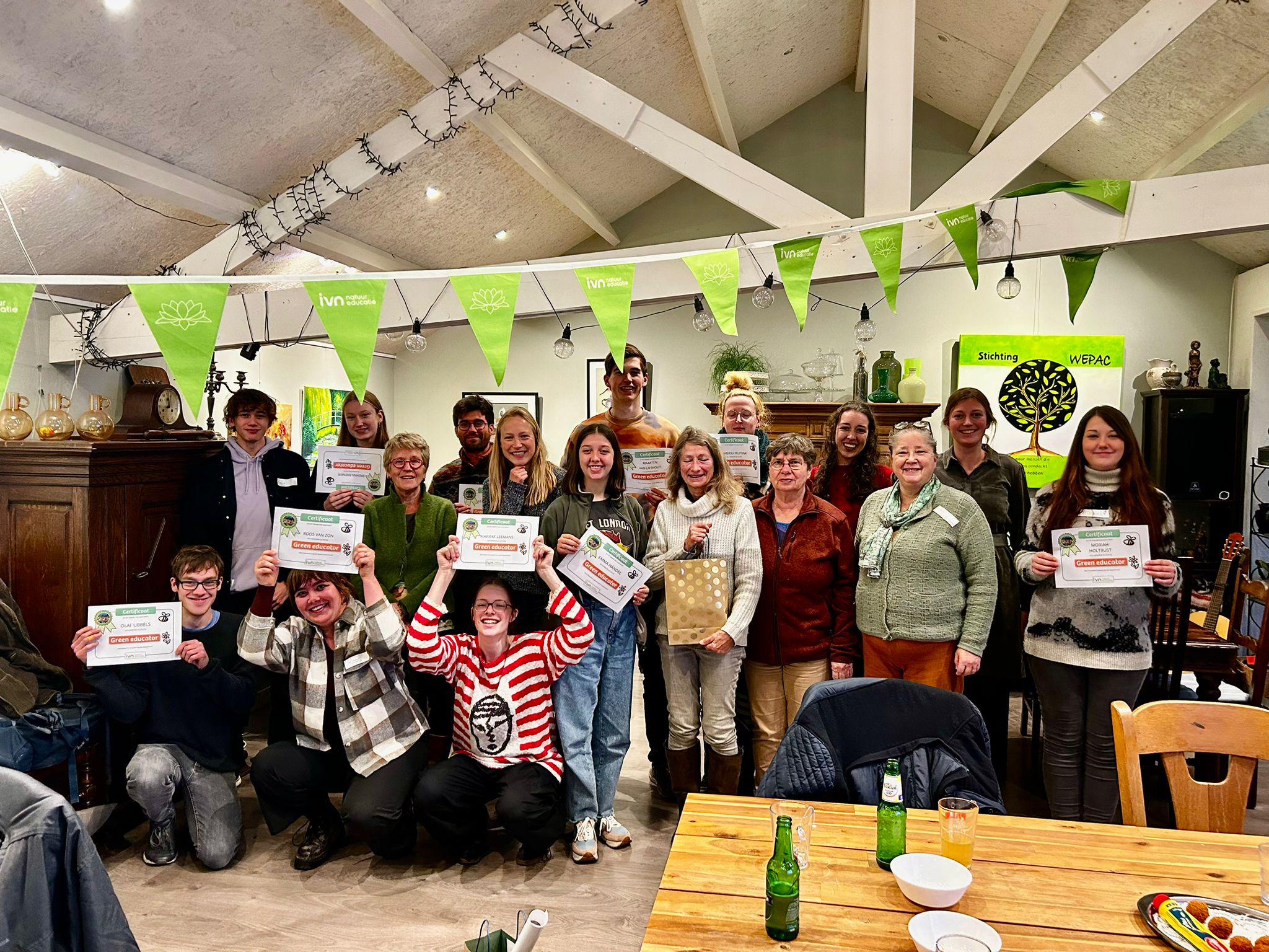 Groep mensen poseert met certificaten in een feestelijke ruimte met groene vlaggetjes.