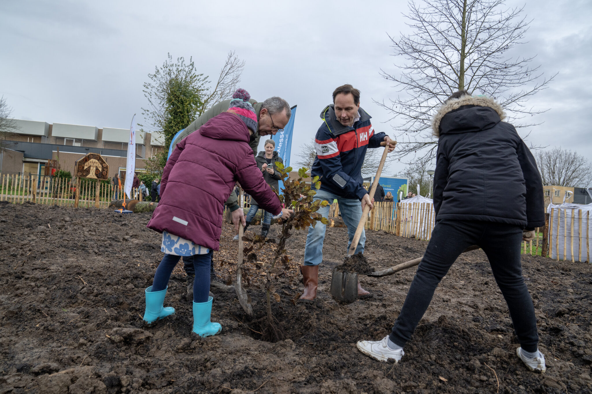 Mensen planten gezamenlijk een boom in een modderig veld omringd door hekken en gebouwen.
