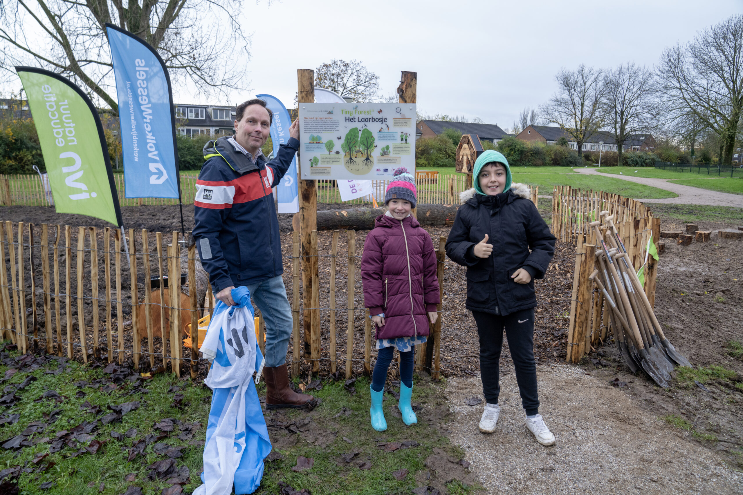 Mensen bij een Tiny Forest bord in een park, met kleurrijke vlaggen en tuingereedschap.