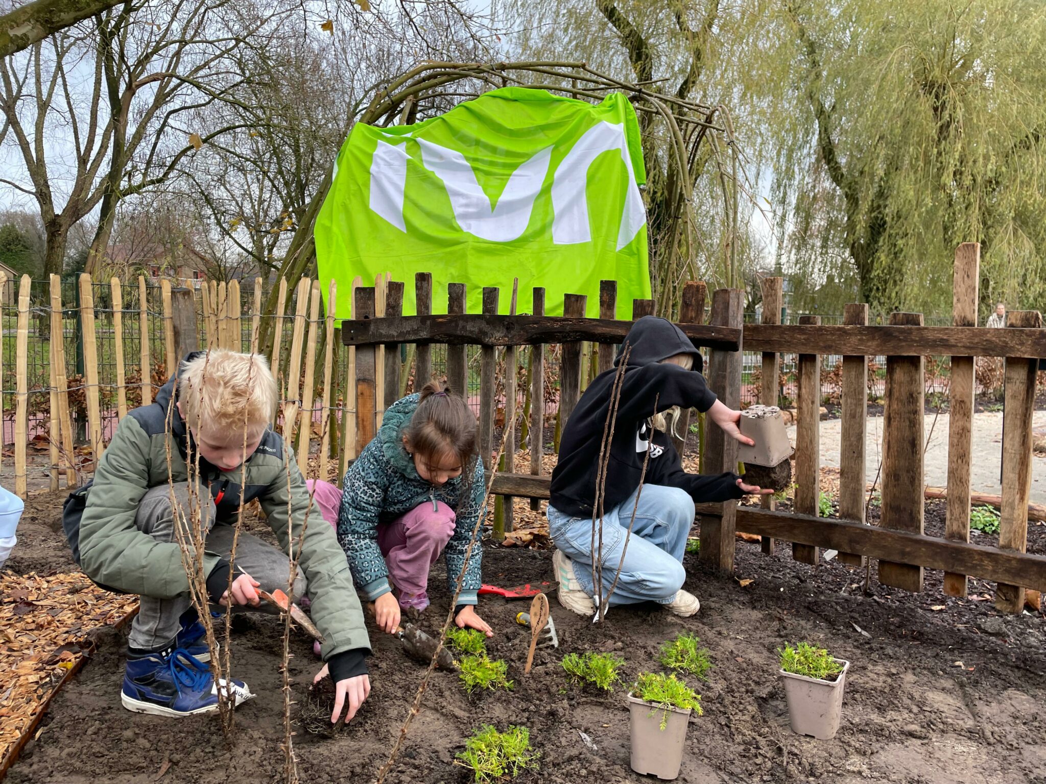 Kinderen planten in een tuin naast een houten hek, groene doek op de achtergrond.