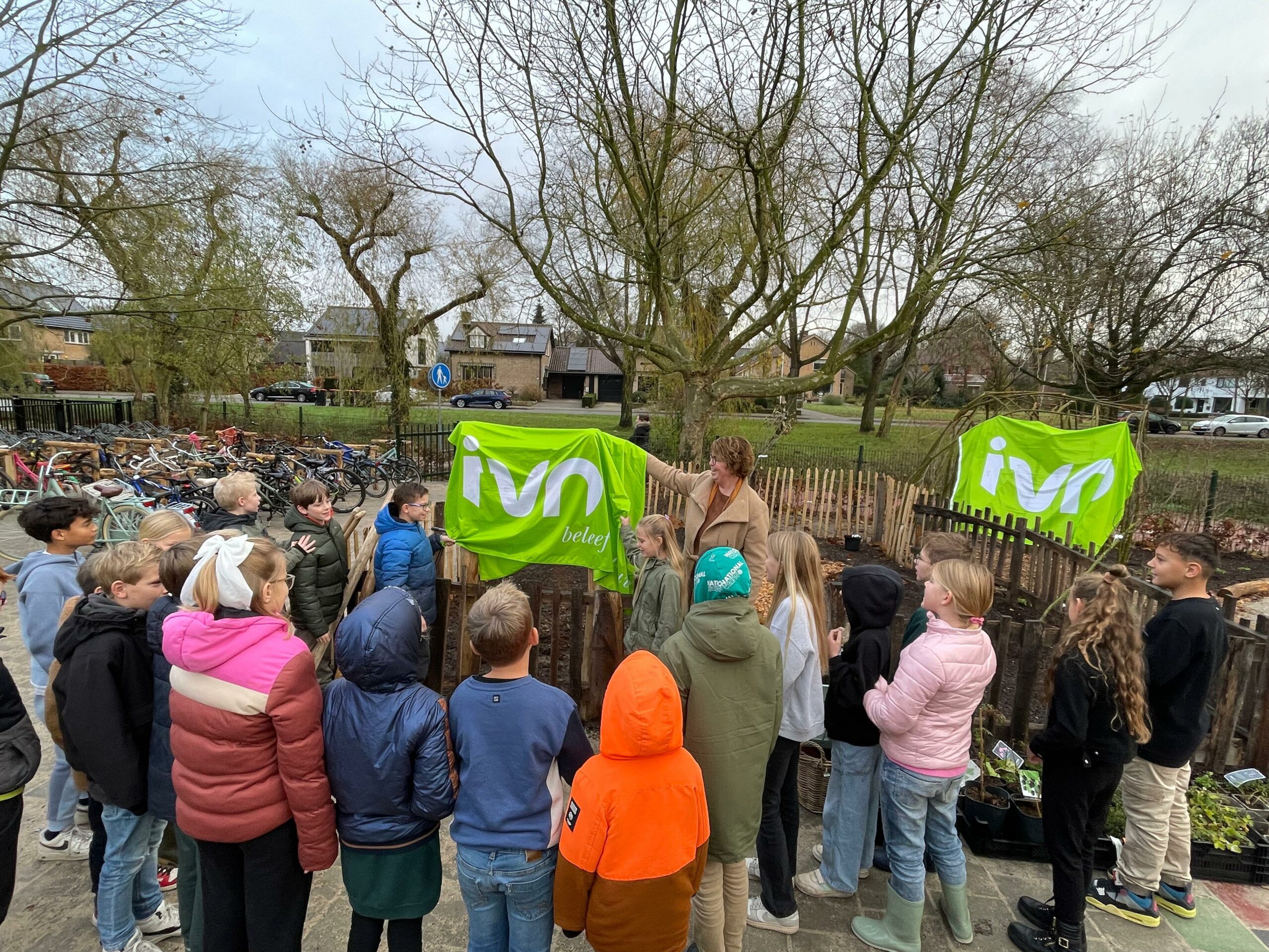 Groep kinderen luistert naar een persoon bij een groene IVN-vlag in een buitenomgeveving met fietsen.