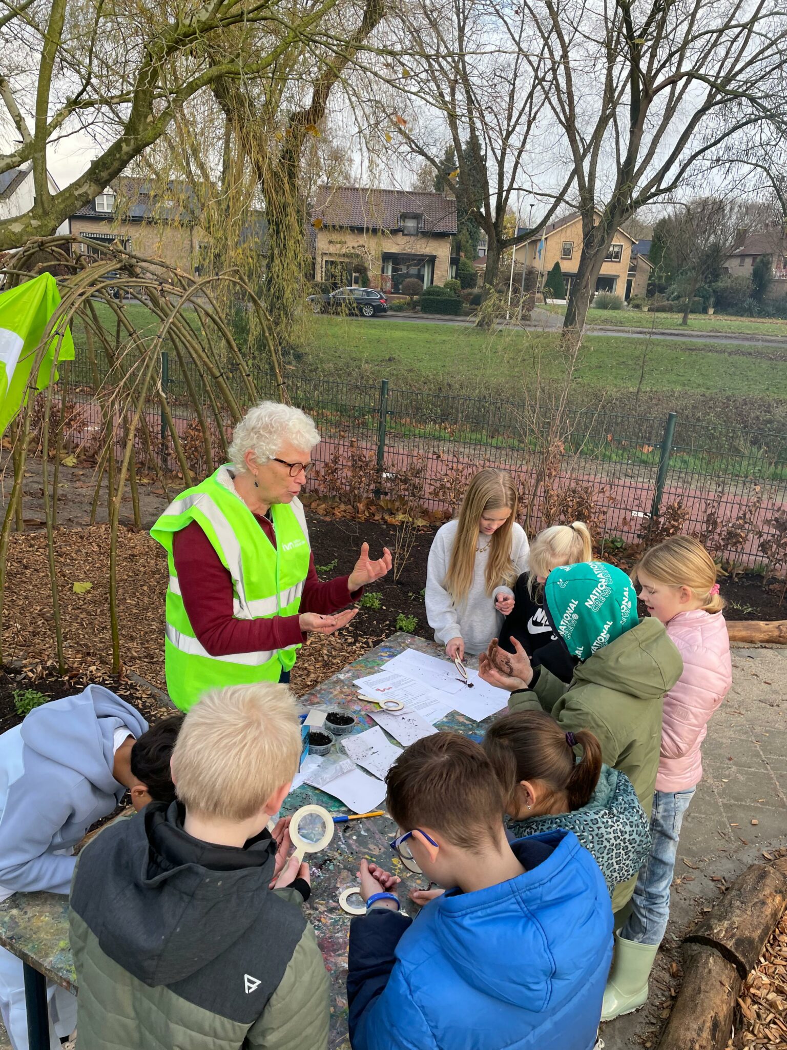 Een volwassene in een gele hes begeleidt kinderen bij een knutselactiviteit buiten.