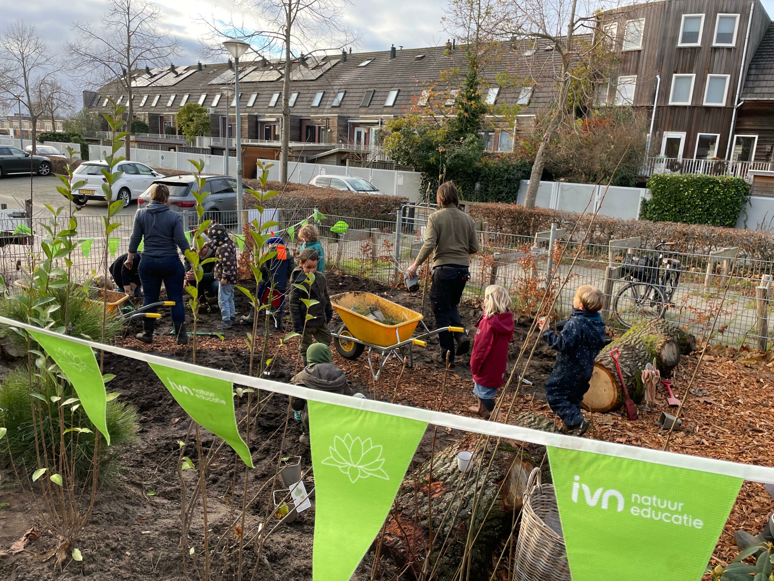 Kinderen planten bomen in een tuin met groene IVN-vlaggen, huizen op de achtergrond.